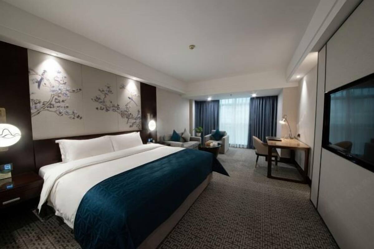 Une chambre d’hôtel avec un lit, un bureau, une chaise, une lampe et une grande fenêtre.