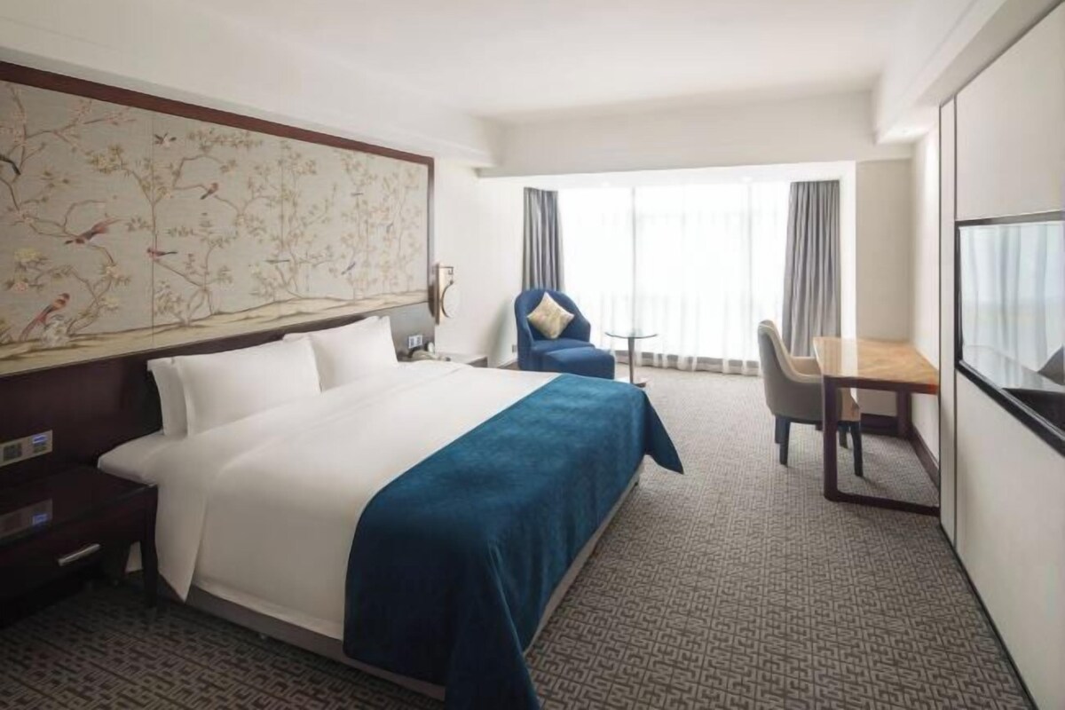 A hotel room with a large bed, a desk, a chair, and a view of the outdoors.