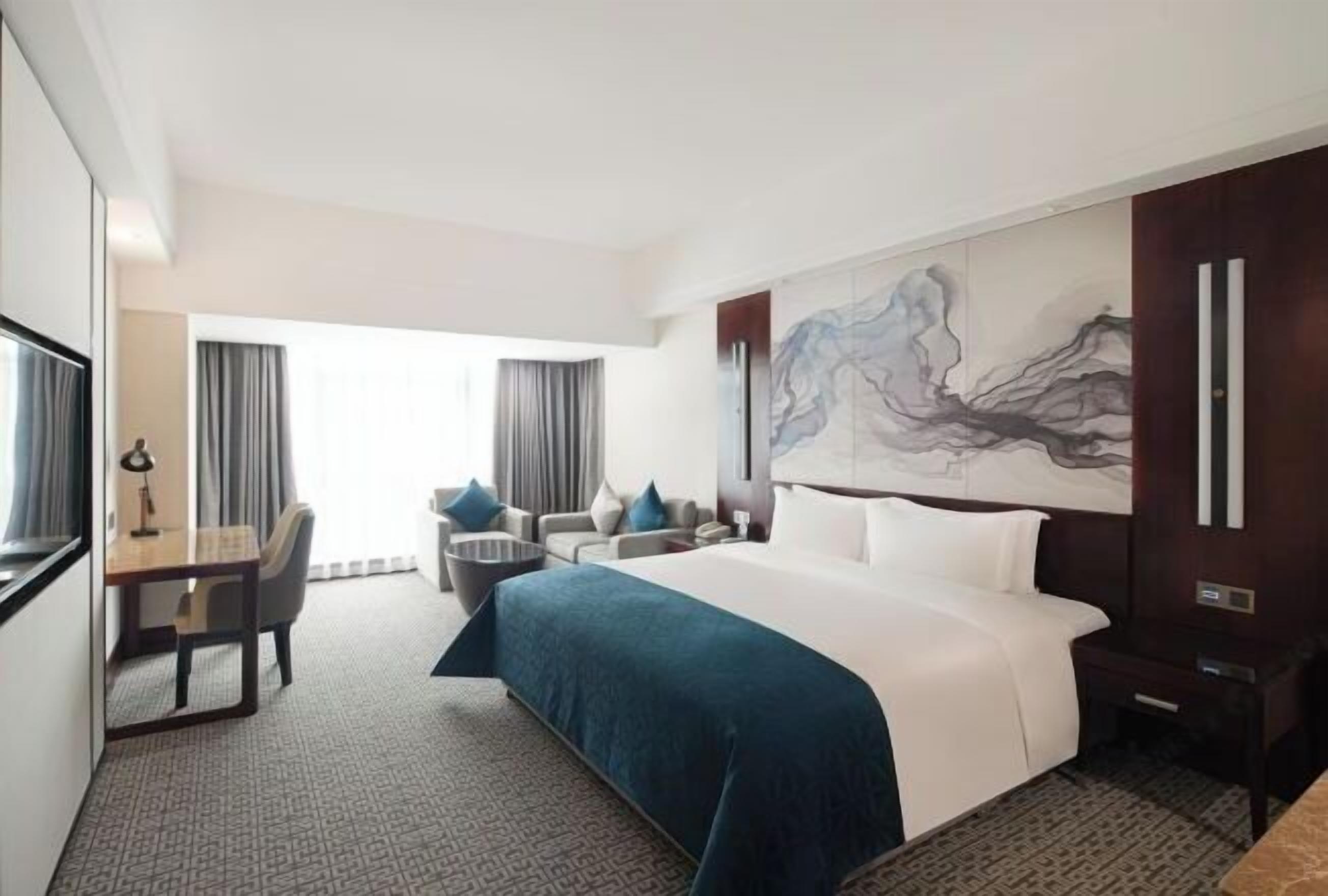 A hotel room with a large bed, a desk with a chair, a sofa, and a large abstract painting on the wall.
