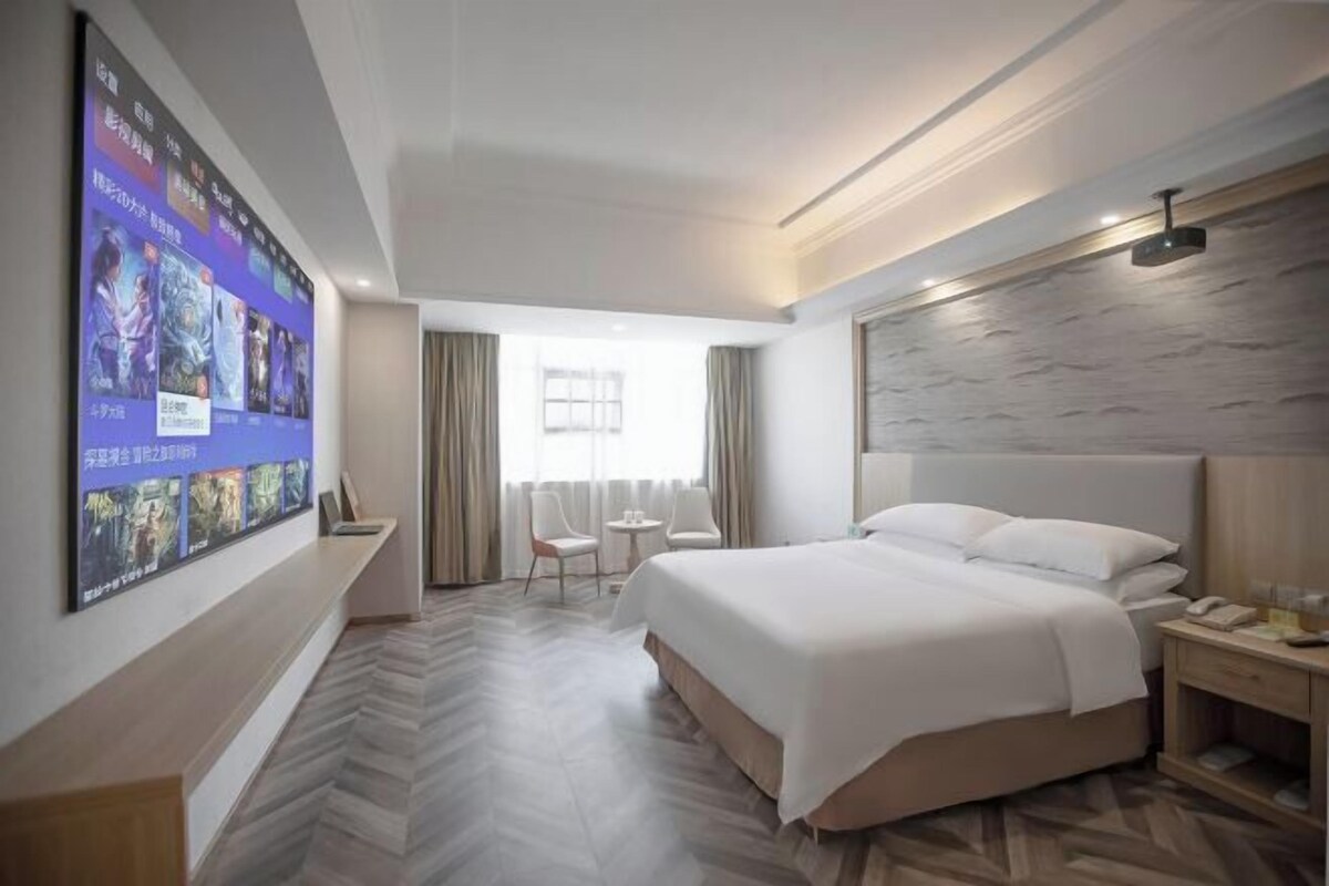 A hotel room with a large bed, a desk, a chair, a small table, and a wall-mounted TV.