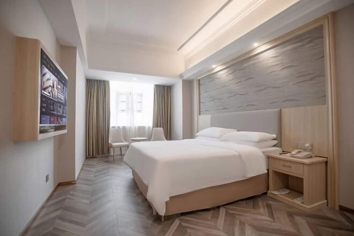 A modern hotel room with a large bed, a bedside table, a chair, and a wall-mounted TV.