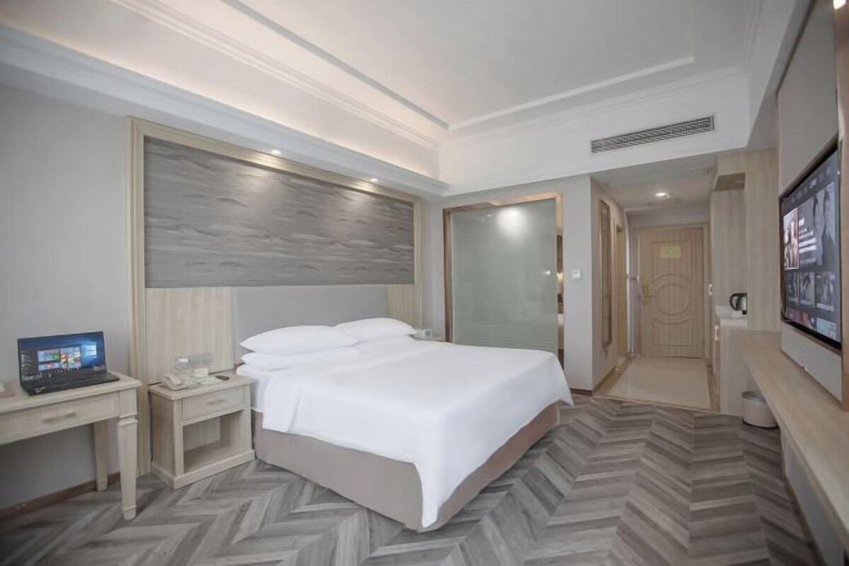A hotel room with a bed, a desk, a laptop, a TV, and a door leading to another room.