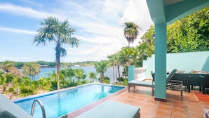 Ein Schwimmbecken mit Meerblick, umgeben von Palmen und einer überdachten Terrasse mit Sitzgelegenheiten.