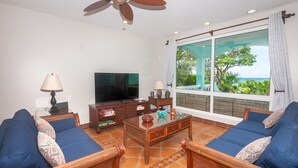 Ein Wohnzimmer mit Fernseher, zwei blauen Sofas, einem Holztisch und einem großen Fenster mit Blick auf Grünflächen und das Meer.