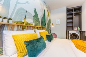 A neatly made bed with yellow and green pillows, a wooden headboard, and a wall mural of tropical plants.