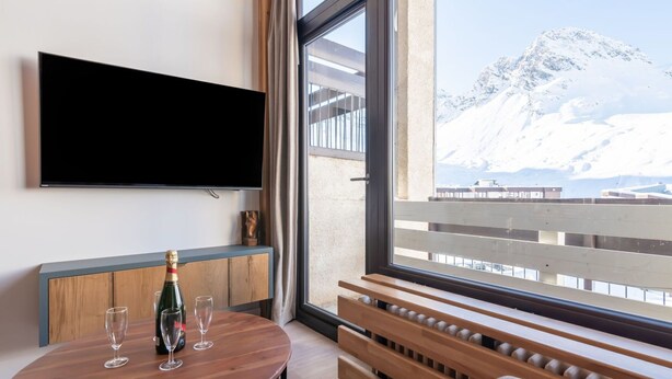 A room with a wooden table, a bottle of champagne, and two glasses. A flat-screen TV is mounted on the wall. The room has a large window with a view of a snowy mountain.