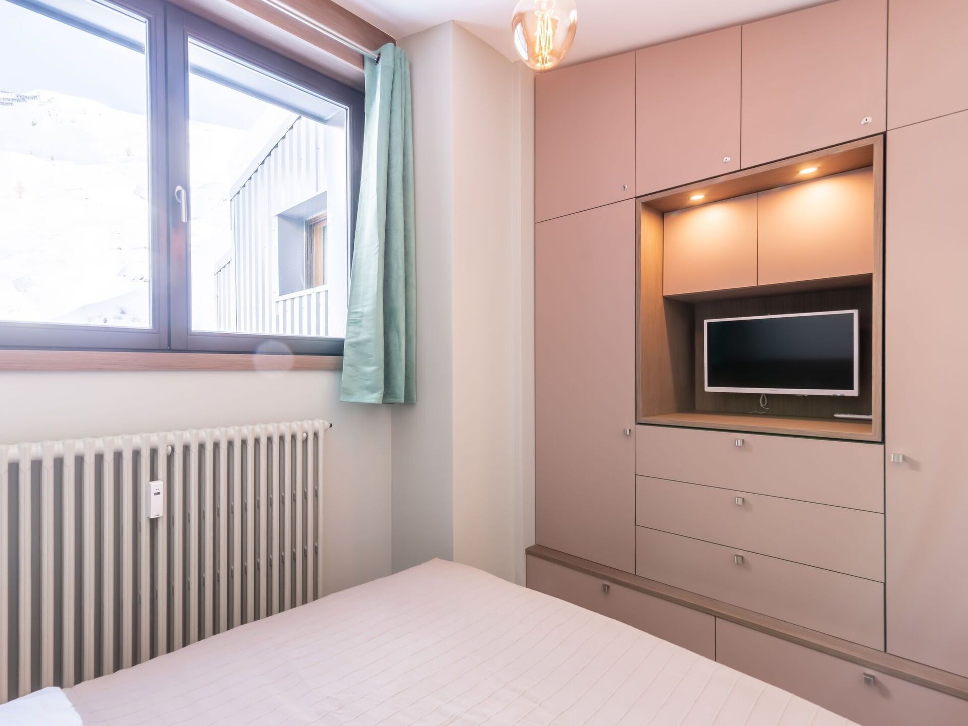 A bedroom with a bed, a window with curtains, a radiator, and built-in TV and storage cabinets.