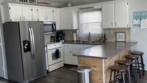 A modern kitchen with white cabinets, stainless steel appliances, a wooden island with stools, and a ceiling fan. - FENCED IN 2nd Row 2 the sea❤️ Shibumi, beach chairs, and linens included. (Surf City)