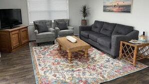 A living room with a grey sofa, two armchairs, a television on a wooden cabinet, a colorful rug, and a painting of birds. - FENCED IN 2nd Row 2 the sea❤️ Shibumi, beach chairs, and linens included. (Surf City)