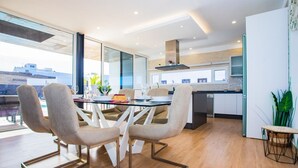 Un espace repas moderne avec une table en verre et des chaises contemporaines, une cuisine en arrière-plan et une vue sur les bâtiments à l’extérieur.
