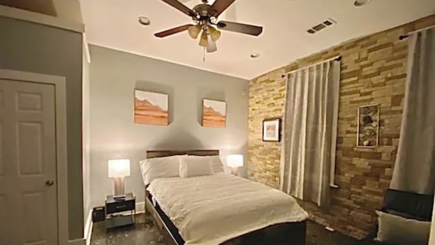 A bedroom with a bed, a ceiling fan, a nightstand, two wall-mounted lamps, and a stone-patterned wall. - Stylish 1 bedroom studio with kitchenette (Jackson)