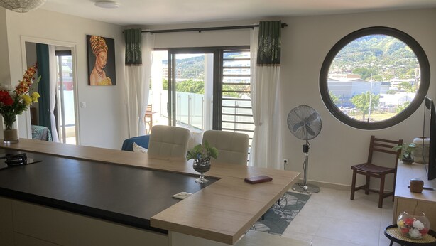 Eine moderne Küche mit einer zentralen Kochinsel, einem Essbereich mit Stühlen, einem runden Tisch und Blick auf eine Stadtlandschaft durch ein Fenster.