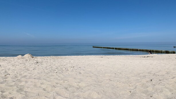 Une plage de sable avec une jetée en bois qui s’avance dans des eaux calmes, sous un ciel bleu dégagé.
