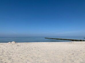 Ein sandiger Strand mit einem hölzernen Steg, der sich in ruhiges Wasser unter blauem Himmel erstreckt.