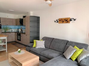 Un salon moderne avec un canapé gris, une table basse en bois et une cuisine en arrière-plan.