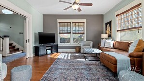 Un salon avec un ventilateur de plafond, un téléviseur, un canapé marron, un tapis à motifs et une fenêtre avec des stores.