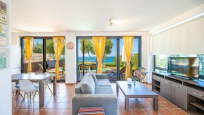 A modern living room with a sofa, dining table, chairs, and a TV. It features large windows with curtains, a view of the sea, and a pool. - Villa Kalma Ena, charming villa, private pool, near Moudania (Gremia Nea Moudania)