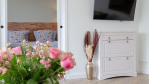 A modern room with a wooden floor, a white cabinet, a flat-screen TV, a round wooden table with two white cups, and a vase with pink flowers.
