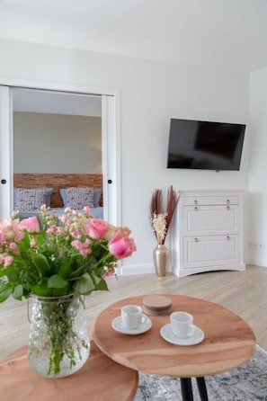 Une chambre moderne avec un plancher en bois, une armoire blanche, un téléviseur à écran plat, une table ronde en bois avec deux tasses blanches et un vase rempli de fleurs roses.