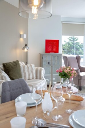 Un coin repas avec une table en bois dressée pour le repas, un canapé avec des coussins à rayures, un meuble avec une enseigne rouge et une fenêtre donnant sur des arbres.