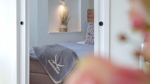 A bedroom with a bed, a bedside table, a shelf with decorative items, and a door leading to another room.