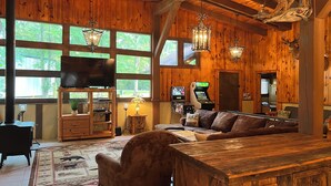 A rustic interior with wooden walls, a large window, a television, a gaming console, and a cozy seating area. - Big Sam Hangout!! 4 bed 3.5 bath South End with rear privacy & Hot Tub (Brookeland)