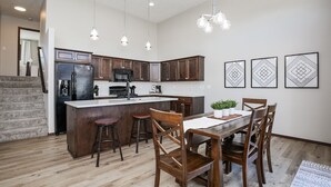 Eine moderne Küche mit dunklen Holdekor-Schränken, einem Edelstahl-Kühlschrank, einer Mikrowelle und einer großen Kochinsel mit Hockern. Über der Kochinsel befinden sich Pendelleuchten und drei gerahmte Wandbilder mit geometrischen Formen.