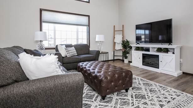 Ein Wohnzimmer mit grauem Sofa, braunem Hocker, weißem Kamin und einem Fernseher auf einem weißen Schrank.
