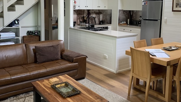 A spacious living area with a brown leather sofa, a wooden coffee table, and a dining table with chairs. A kitchen with white cabinets and a stainless steel refrigerator is visible in the background. - TreeTops - wood fire, outdoor spa and great coffee (Hepburn Springs)