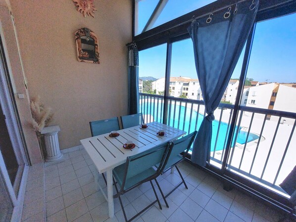 Een balkon met een tafel en stoelen, uitzicht op een zwembad en een gebouw op de achtergrond.