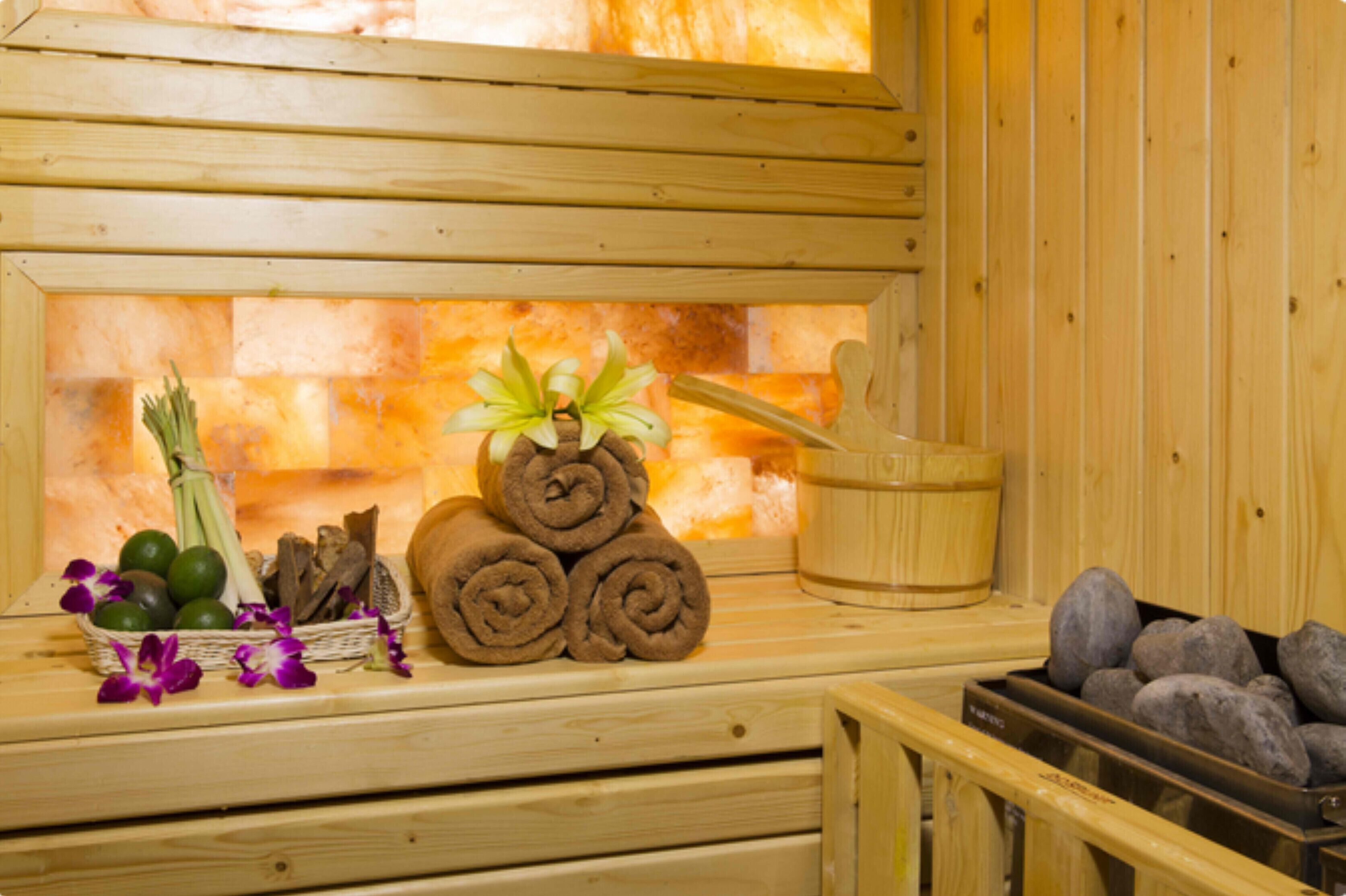 A wooden sauna with a stone wall, a wooden bench, a basket of towels, a bucket, and a tray of stones.