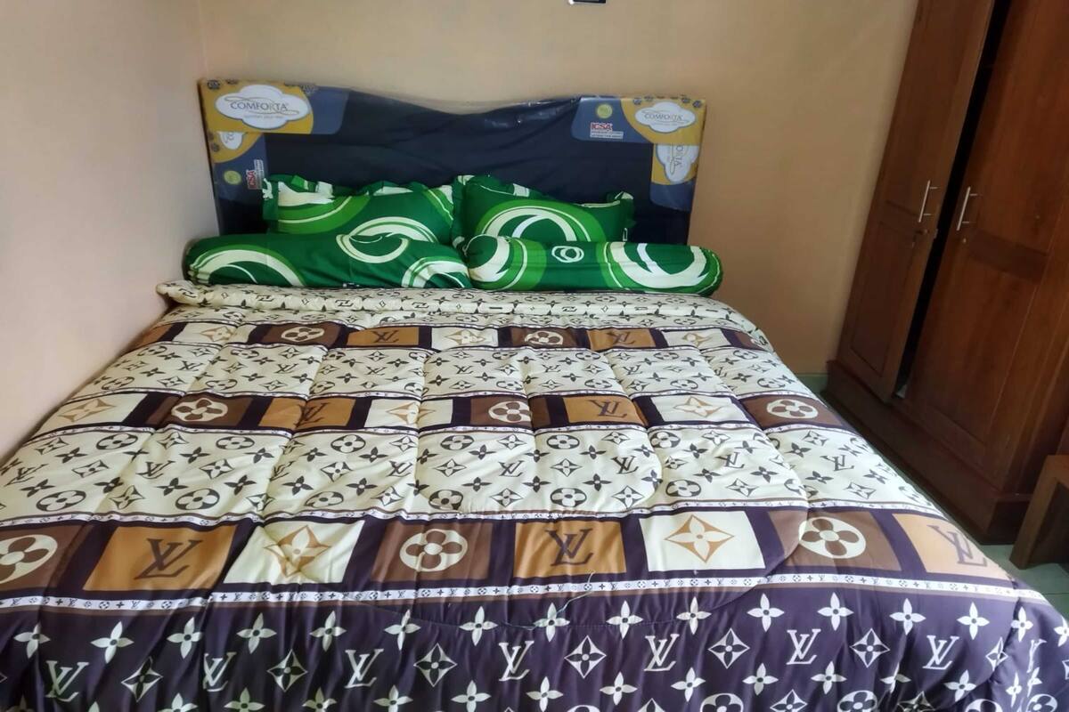 Un lit avec une couvre-lit à motifs et des oreillers, une armoire en bois et un petit tabouret en bois.