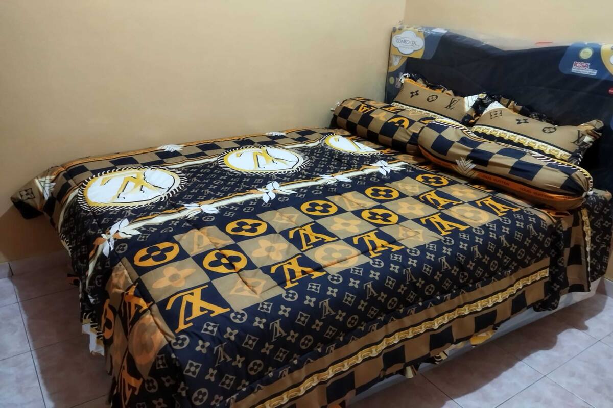 Un lit avec une couvre-lit à motifs et des coussins assortis, une tête de lit ornée d’un logo et une étagère fixée au mur.