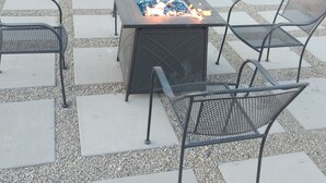 Een terras buiten met een vierkante vuurkorf, omringd door metalen stoelen en een tafel.