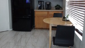 Een kleine keuken met houten kastjes, een zwarte koelkast, een koffiezetapparaat en een eettafel met twee stoelen.