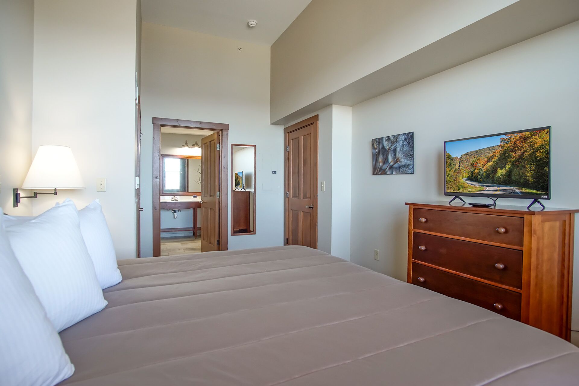 A bedroom with a bed, a dresser, a television, and a bathroom visible through an open door.