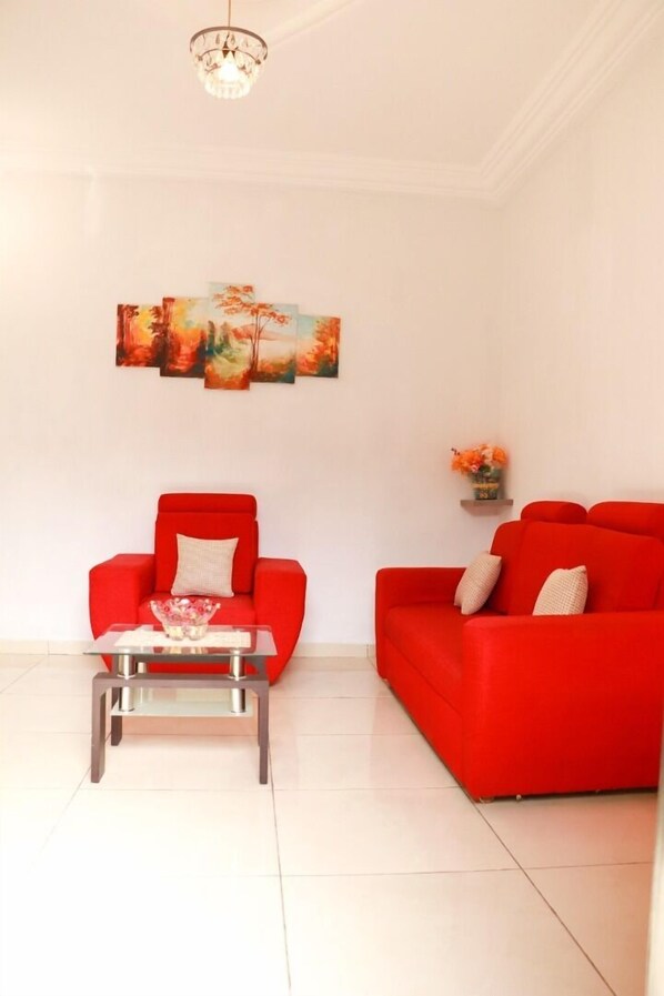 Un salon avec un canapé rouge, un fauteuil rouge et une table basse en verre. Un lustre est suspendu au plafond et un tableau orne le mur.
