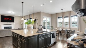 A modern kitchen with a large island, dark cabinetry, and a dining area with a view. - Sunset Casa: Golf & Lake Views, Hot Tub, Fire Pit, Sleeps 16 (Aurora)