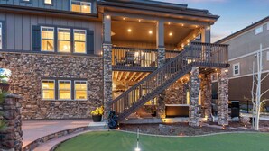 A two-story house with a stone facade, a covered porch, and a backyard with a putting green. - Sunset Casa: Golf & Lake Views, Hot Tub, Fire Pit, Sleeps 16 (Aurora)