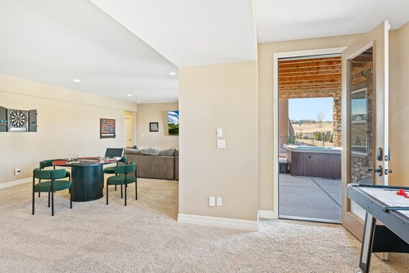 A living room with a dartboard, a table with chairs, a couch, and a view of an outdoor patio with a hot tub.