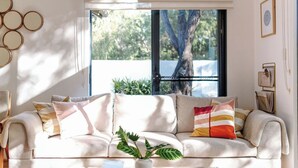 A modern living room with a beige sofa, a glass coffee table, and a large window with a view of trees and a fence. - The Villa | Margaret River (Margaret River)