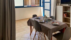 Un comedor con una mesa lista para cuatro personas, un televisor sobre un mueble bajo y una estantería con libros y objetos decorativos.