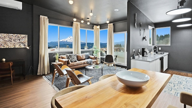 Una sala de estar moderna con un ventanal grande, un comedor con mesa de madera y una zona de cocina con lavamanos y encimera.