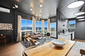 Un salon moderne doté d’une grande fenêtre, un coin repas avec une table en bois et un espace cuisine avec un évier et un plan de travail.