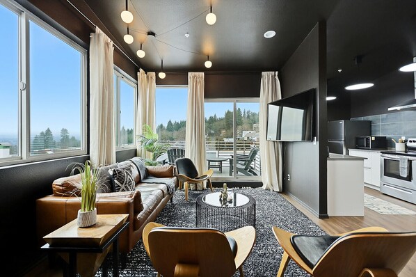 Un salon moderne avec un canapé, des fauteuils, une table basse et un téléviseur. Il y a un balcon avec vue sur les arbres et les bâtiments.