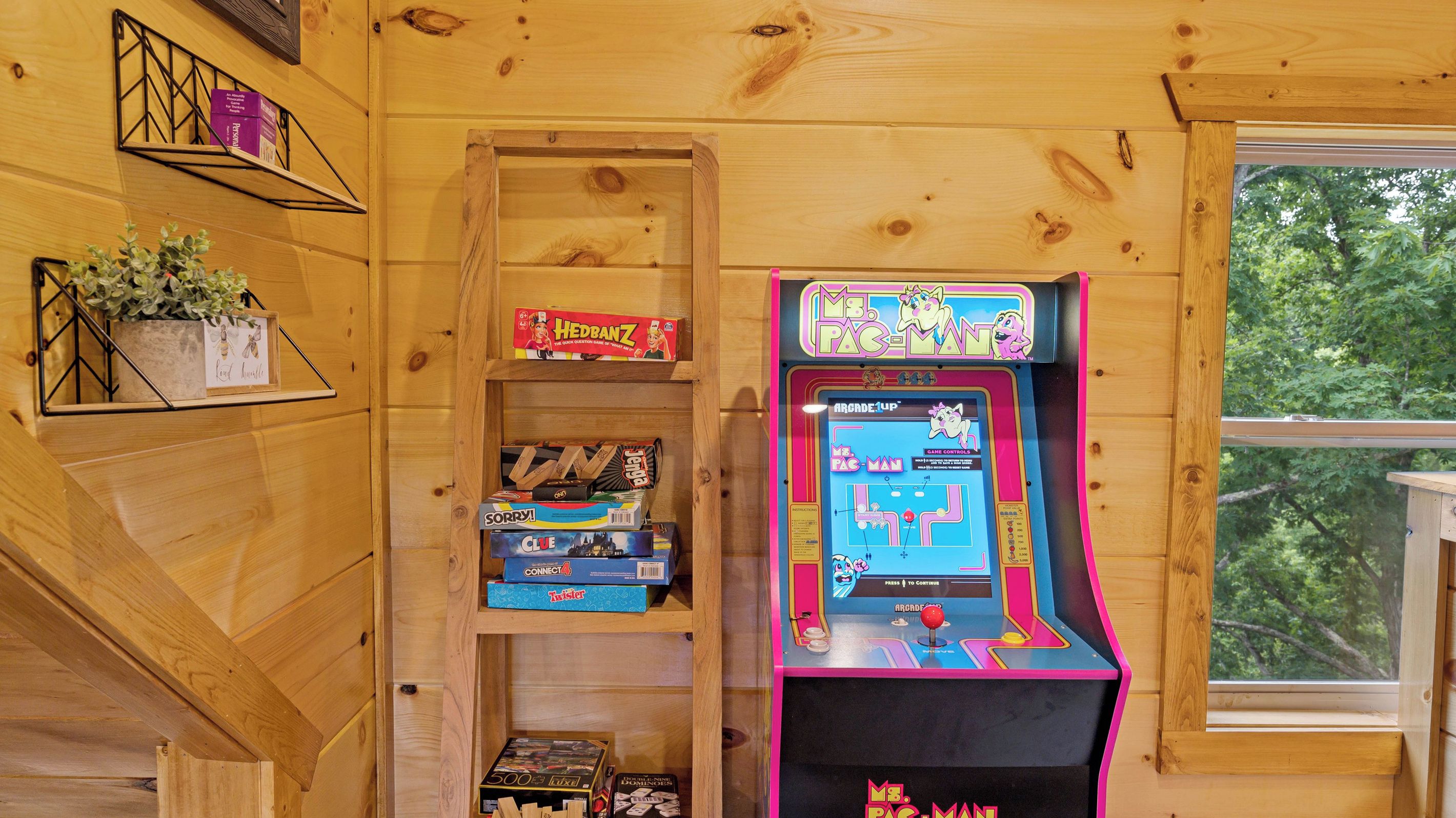 Un interior de madera con una máquina de videojuegos Pac-Man, estantes con juegos de mesa y un ventanal con vista a árboles.