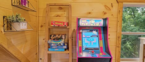 A wooden interior with a Pac-Man arcade machine, shelves with board games, and a window with a view of trees.