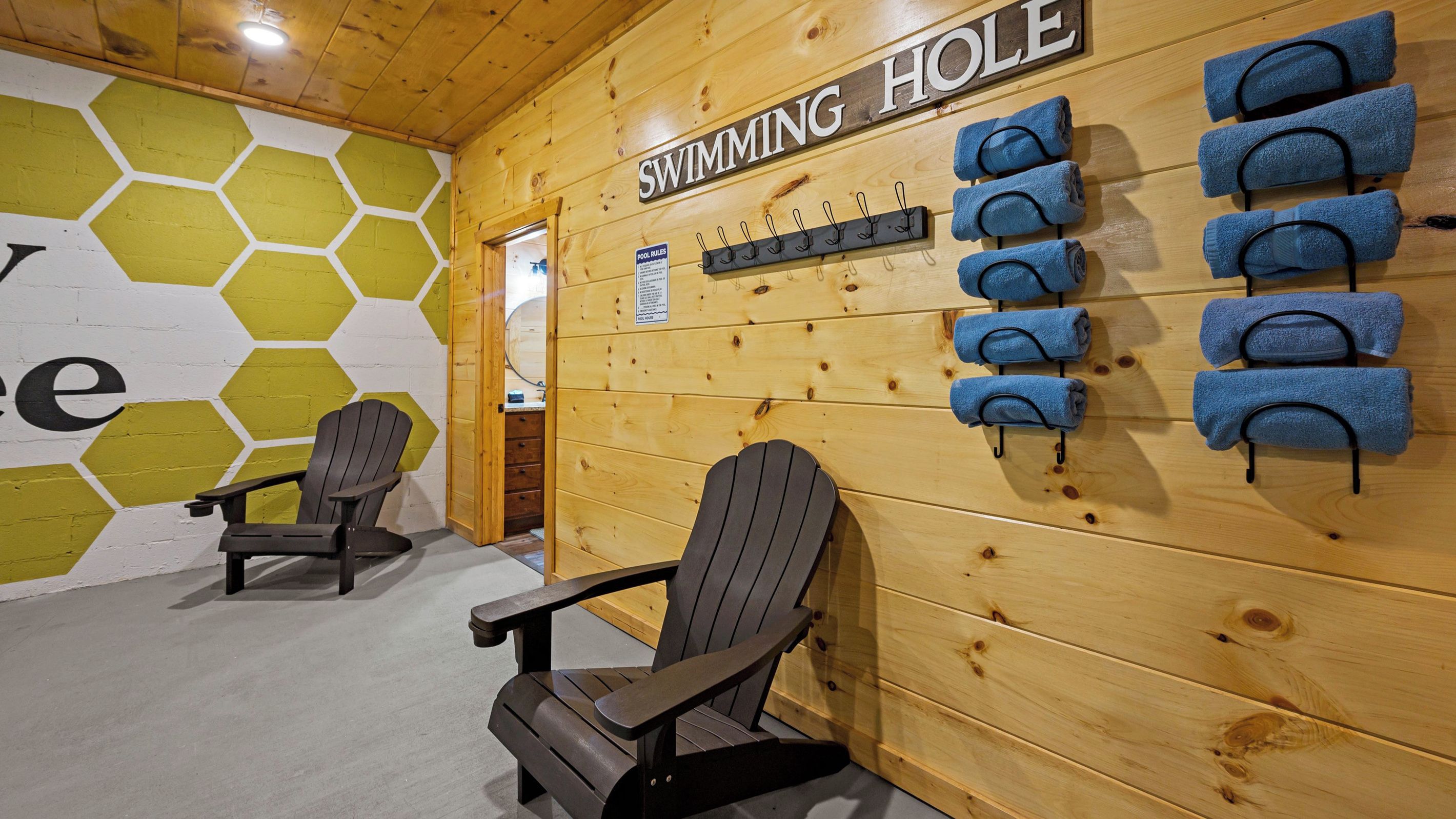 A wooden interior with a sign reading 'SWIMMING HOLE,' a wall-mounted towel rack with towels, and two Adirondack chairs.
