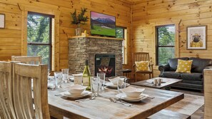 A wooden cabin interior with a stone fireplace, dining table set for four, and a leather sofa.