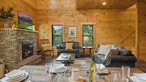 A cozy cabin interior with a stone fireplace, wooden walls, a leather sofa, and a dining table set for two.